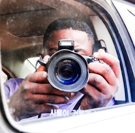 A person is holding a DSLR camera, capturing an image through the side mirror of a vehicle. The focus is on the camera lens, and the person's hands are visible. The background shows the interior of the car and a reflection.