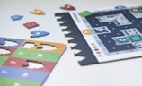 A close-up view of a strategic board game set on a light surface. The scene includes game pieces that resemble arrows and a board with a grid-like layout marked by letters and numbers. Colored pieces, mostly in blue, red, green, and yellow, are scattered around. The board has icons and symbols suggesting it might involve navigation or directional tasks.