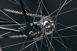 A close-up view of a bicycle wheel focusing on the hub and spokes. The metallic spokes radiate from the central hub, which is intricately designed and features visible screws and a rotating mechanism. The chain and part of the frame are also visible, demonstrating precision engineering and craftsmanship.