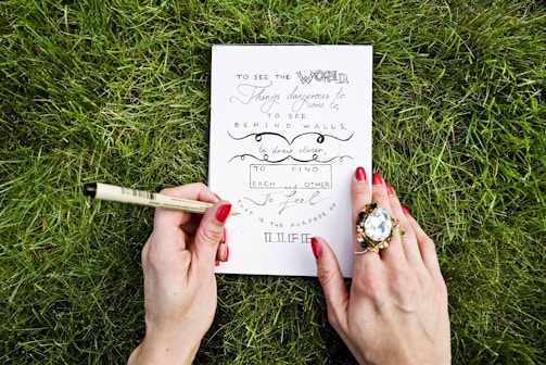 A person is writing with a pen on a piece of paper that contains an inspirational quote, surrounded by lush green grass. The person's nails are painted red, and they wear a large, ornate ring on one hand.