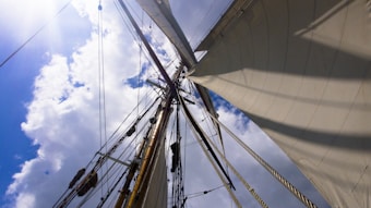 View looking up at a sailboat's mast and sails, capturing the rigging and ropes against a partly cloudy sky with sunlight peeking through.