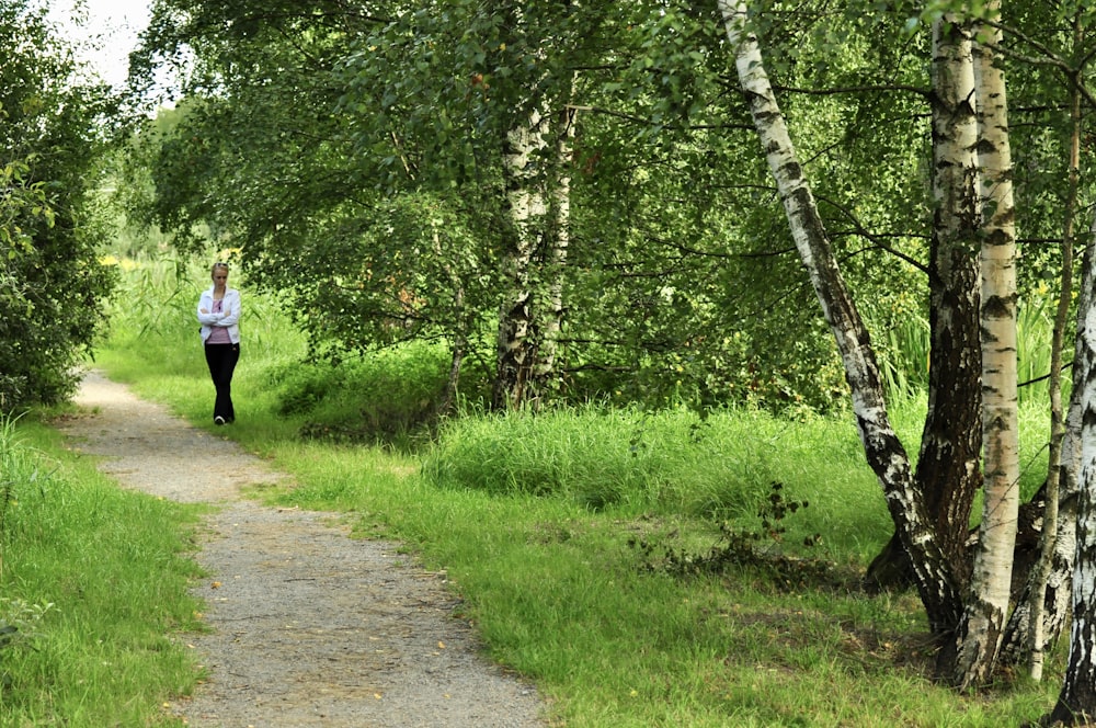 mujer con camisa blanca de manga larga y pantalones negros caminando por el sendero entre la hierba verde y