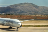 An Emirates airplane is on a runway, set against a backdrop of a large mountain and a few scattered trees. The aircraft is mostly white with the airline's branding prominently displayed on its side. The scene appears to be part of an airport, with the runway and taxiway visible in the foreground.
