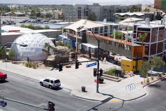 An urban setting features a modern architectural complex with a dome structure and a container-style building. The area includes greenery and a few sculptures, suggesting an artistic atmosphere. A sign indicates it is 'Container Park.' Streets and parked cars are visible, and a few pedestrians walk nearby.