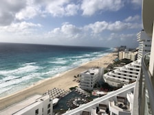 A beachfront scene with white sandy shores and vibrant blue ocean waves brushing against the coast. High-rise buildings with balconies line the shoreline, and in the foreground, there are pools and sunbathing areas with umbrellas. The sky is partly cloudy, casting some shadow and light over the entire view.