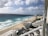 A beachfront scene with white sandy shores and vibrant blue ocean waves brushing against the coast. High-rise buildings with balconies line the shoreline, and in the foreground, there are pools and sunbathing areas with umbrellas. The sky is partly cloudy, casting some shadow and light over the entire view.