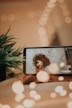 A smartphone placed on a table displays an image of a woman with curly hair, wearing glasses, looking sideways. The background features a decorative wall. In the foreground, blurred fairy lights create a warm bokeh effect, adding an artistic touch. A green plant decorates the scene.