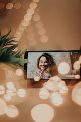 A phone on a table displays a smiling person with glasses on its screen. In the foreground and background, there are decorative bokeh lights creating a warm ambiance. A plant with green leaves is slightly visible on the left side.