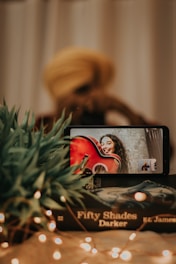 A smartphone displays a video call with a person holding a red guitar and smiling. In the foreground, a stack of books titled 'Fifty Shades Darker' and some green plants are visible. Fairy lights add a warm, cozy ambiance to the scene.