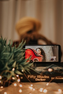 A smartphone displays a video call with a person holding a red guitar and smiling. In the foreground, a stack of books titled 'Fifty Shades Darker' and some green plants are visible. Fairy lights add a warm, cozy ambiance to the scene.