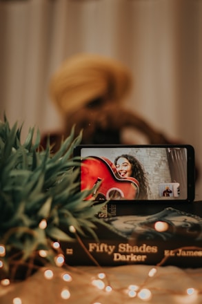 A smartphone displays a video call with a person holding a red guitar and smiling. In the foreground, a stack of books titled 'Fifty Shades Darker' and some green plants are visible. Fairy lights add a warm, cozy ambiance to the scene.