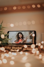 A smartphone displays a video call with a woman holding a guitar, smiling warmly against a softly blurred background. The scene is illuminated by warm, bokeh fairy lights, adding a cozy, intimate atmosphere. Nearby, a book titled 'Fifty Shades Darker' is partially visible, enhancing the setting.