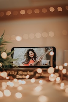 A smartphone displays a video call with a woman holding a guitar, smiling warmly against a softly blurred background. The scene is illuminated by warm, bokeh fairy lights, adding a cozy, intimate atmosphere. Nearby, a book titled 'Fifty Shades Darker' is partially visible, enhancing the setting.