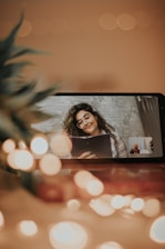 A smartphone displaying a video call screen where a woman with glasses and curly hair holds a clipboard, surrounded by soft bokeh lights and a blurred plant in the foreground. The setting appears cozy with warm tones.