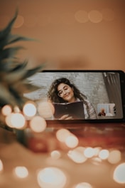 A smartphone displaying a video call screen where a woman with glasses and curly hair holds a clipboard, surrounded by soft bokeh lights and a blurred plant in the foreground. The setting appears cozy with warm tones.