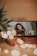 A smartphone displaying a video call screen where a woman with glasses and curly hair holds a clipboard, surrounded by soft bokeh lights and a blurred plant in the foreground. The setting appears cozy with warm tones.