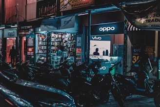A busy street scene at night with several parked motorcycles in the foreground. The background features brightly lit retail shops, including a store displaying Oppo and Vivo signs. The shop’s well-lit interiors showcase mobile phone accessories, and a few people are visible inside and around the shop.