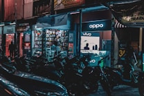 A busy street scene at night with several parked motorcycles in the foreground. The background features brightly lit retail shops, including a store displaying Oppo and Vivo signs. The shop’s well-lit interiors showcase mobile phone accessories, and a few people are visible inside and around the shop.