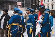A group of uniformed men in blue military dress with ceremonial decorations are standing and conversing. One man is holding a flag, and other individuals and buildings are in the background.