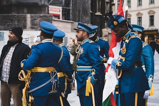 A group of uniformed men in blue military dress with ceremonial decorations are standing and conversing. One man is holding a flag, and other individuals and buildings are in the background.
