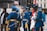 A group of uniformed men in blue military dress with ceremonial decorations are standing and conversing. One man is holding a flag, and other individuals and buildings are in the background.