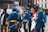 A group of uniformed men in blue military dress with ceremonial decorations are standing and conversing. One man is holding a flag, and other individuals and buildings are in the background.