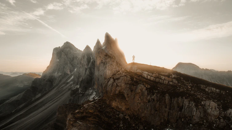 A lone trail runner ascending a rugged mountain ridge at dawn, with dramatic shadows and a fiery orange sky.