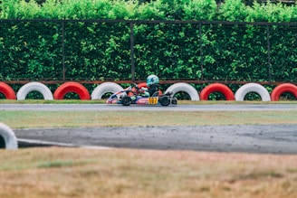 A go-kart is racing on a track with a driver wearing a helmet. The track is lined with alternating red and white tire barriers, and a lush green hedge serves as a backdrop.