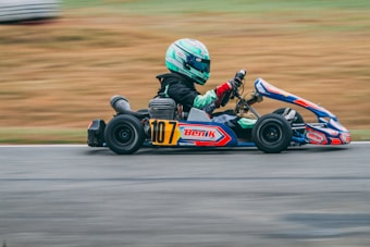 A person wearing a green helmet and black race suit drives a go-kart on a racetrack. The go-kart is blue with red and white accents, featuring the number 107 prominently. The background is blurred, indicating motion and speed.