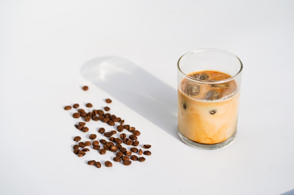 A minimalist composition featuring a glass of iced coffee with a creamy, swirling texture set against a clean white background. Scattered coffee beans on the left add an element of texture and interest, with their rich brown color contrasting the lighter tones of the beverage.