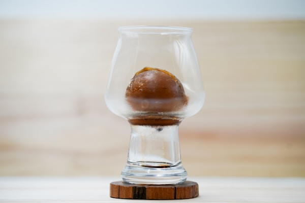 A clear glass sits on a wooden coaster, containing a round, brown object that appears to be ice. The background is softly blurred with warm, neutral tones.