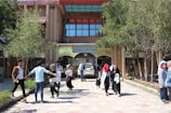 A bustling scene in front of a building entrance with several people walking and interacting. The entrance features an Arabic sign above the doorway. Trees and shrubs surround the pavement, adding greenery to the area. Some individuals are carrying books or bags, suggesting a school or university setting.