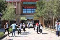 A bustling scene in front of a building entrance with several people walking and interacting. The entrance features an Arabic sign above the doorway. Trees and shrubs surround the pavement, adding greenery to the area. Some individuals are carrying books or bags, suggesting a school or university setting.