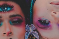 Two faces adorned with vibrant makeup and surreal elements are juxtaposed, one with an additional eye painted on the forehead. The makeup features bright shades of blue, pink, and purple, with ornate details including rhinestones and a butterfly accessory. The artwork blends reality and fantasy, creating a striking visual impact.