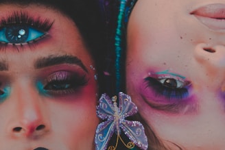 Two faces adorned with vibrant makeup and surreal elements are juxtaposed, one with an additional eye painted on the forehead. The makeup features bright shades of blue, pink, and purple, with ornate details including rhinestones and a butterfly accessory. The artwork blends reality and fantasy, creating a striking visual impact.