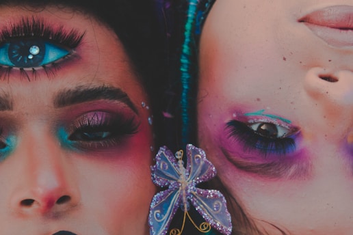 Two faces adorned with vibrant makeup and surreal elements are juxtaposed, one with an additional eye painted on the forehead. The makeup features bright shades of blue, pink, and purple, with ornate details including rhinestones and a butterfly accessory. The artwork blends reality and fantasy, creating a striking visual impact.