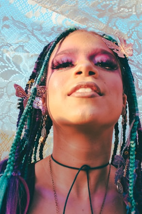 A person with colorful braided hair adorned with butterfly accessories. The individual is wearing dramatic makeup with vibrant eye shadows and long eyelashes. They have multiple earrings and a choker necklace. The background consists of a lace texture with floral designs.