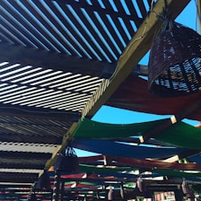 A wooden pergola structure with slatted overhead beams creating a pattern of light and shadows. Colorful fabric panels, in green, blue, and red, are stretched across parts of the pergola. Wicker cone-shaped light fixtures hang from the beams, and the sky is clear and bright blue in the background.