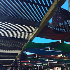 A wooden pergola structure with slatted overhead beams creating a pattern of light and shadows. Colorful fabric panels, in green, blue, and red, are stretched across parts of the pergola. Wicker cone-shaped light fixtures hang from the beams, and the sky is clear and bright blue in the background.