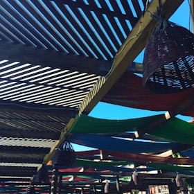 A wooden pergola structure with slatted overhead beams creating a pattern of light and shadows. Colorful fabric panels, in green, blue, and red, are stretched across parts of the pergola. Wicker cone-shaped light fixtures hang from the beams, and the sky is clear and bright blue in the background.