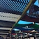 A wooden pergola structure with slatted overhead beams creating a pattern of light and shadows. Colorful fabric panels, in green, blue, and red, are stretched across parts of the pergola. Wicker cone-shaped light fixtures hang from the beams, and the sky is clear and bright blue in the background.