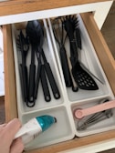 A kitchen drawer with white dividers containing various black plastic kitchen utensils such as spoons, spatulas, and a whisk. An electric hand-held vacuum is being used to clean one of the compartments. Pink measuring spoons and metallic measuring cups are neatly arranged in another compartment.