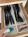 A kitchen drawer with white dividers containing various black plastic kitchen utensils such as spoons, spatulas, and a whisk. An electric hand-held vacuum is being used to clean one of the compartments. Pink measuring spoons and metallic measuring cups are neatly arranged in another compartment.