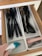 A kitchen drawer with white dividers containing various black plastic kitchen utensils such as spoons, spatulas, and a whisk. An electric hand-held vacuum is being used to clean one of the compartments. Pink measuring spoons and metallic measuring cups are neatly arranged in another compartment.