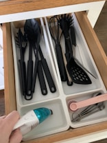 A kitchen drawer with white dividers containing various black plastic kitchen utensils such as spoons, spatulas, and a whisk. An electric hand-held vacuum is being used to clean one of the compartments. Pink measuring spoons and metallic measuring cups are neatly arranged in another compartment.