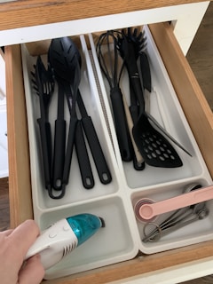 A kitchen drawer with white dividers containing various black plastic kitchen utensils such as spoons, spatulas, and a whisk. An electric hand-held vacuum is being used to clean one of the compartments. Pink measuring spoons and metallic measuring cups are neatly arranged in another compartment.