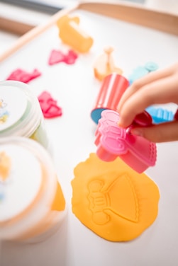 A playful scene featuring a hand pressing a pink mold onto a sheet of orange playdough, creating an imprint of a dress. Around the main activity, various colorful molds and containers are scattered, suggesting a creative, crafting session.