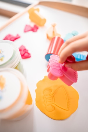 A playful scene featuring a hand pressing a pink mold onto a sheet of orange playdough, creating an imprint of a dress. Around the main activity, various colorful molds and containers are scattered, suggesting a creative, crafting session.