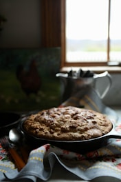 A freshly baked round cake or bread rests in a dark skillet on top of a decorative cloth with floral and geometric patterns. The scene is lit softly from a window in the background, which casts a warm glow over the kitchen area.