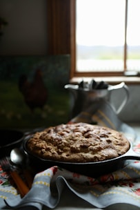 A freshly baked round cake or bread rests in a dark skillet on top of a decorative cloth with floral and geometric patterns. The scene is lit softly from a window in the background, which casts a warm glow over the kitchen area.